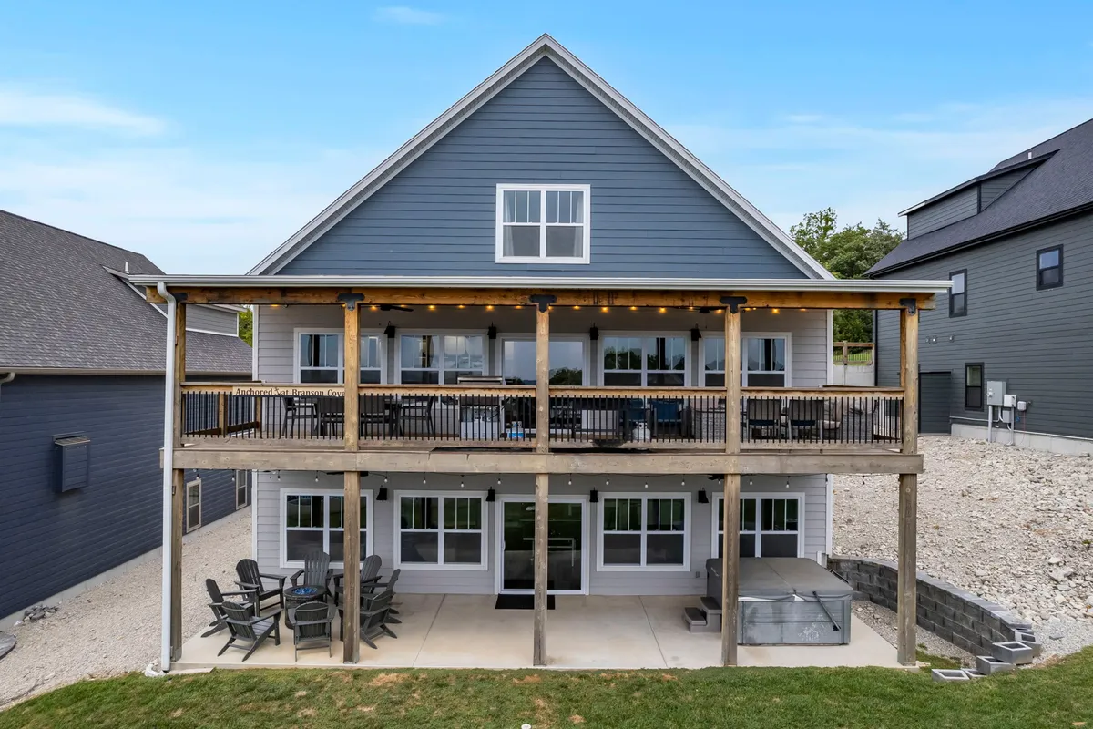 The image features the exterior of a spacious, beautifully designed single-family house in Hollister, Missouri. A large deck with inviting outdoor seating wraps around the home, offering ample space for groups to gather. The symmetrical architecture of the house creates a sense of elegance and welcome. The deck overlooks scenic natural surroundings—perfect for sunset dinners, morning coffee, or stargazing under the open sky.