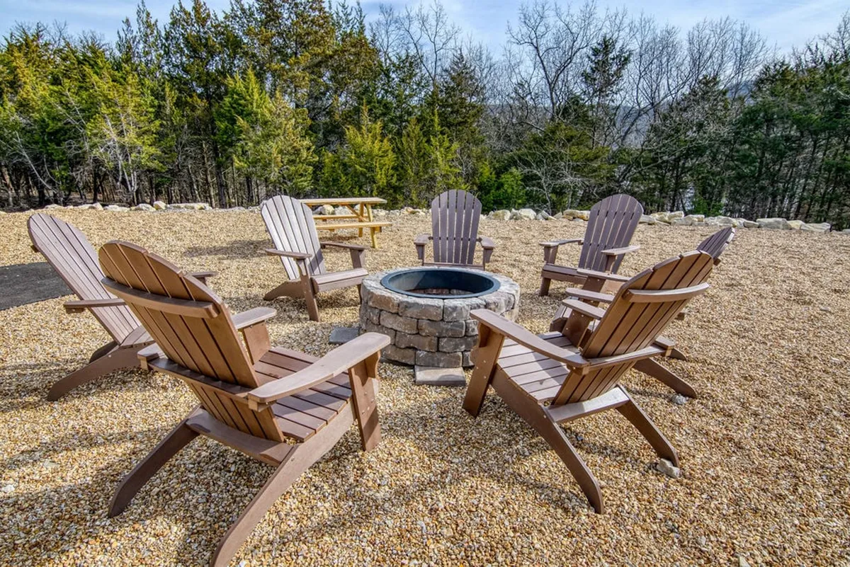 A cozy communal firepit area surrounded by Adirondack chairs, set amidst trees on a gravel ground. The scene suggests a gathering place for socializing, storytelling, and roasting marshmallows under the stars.