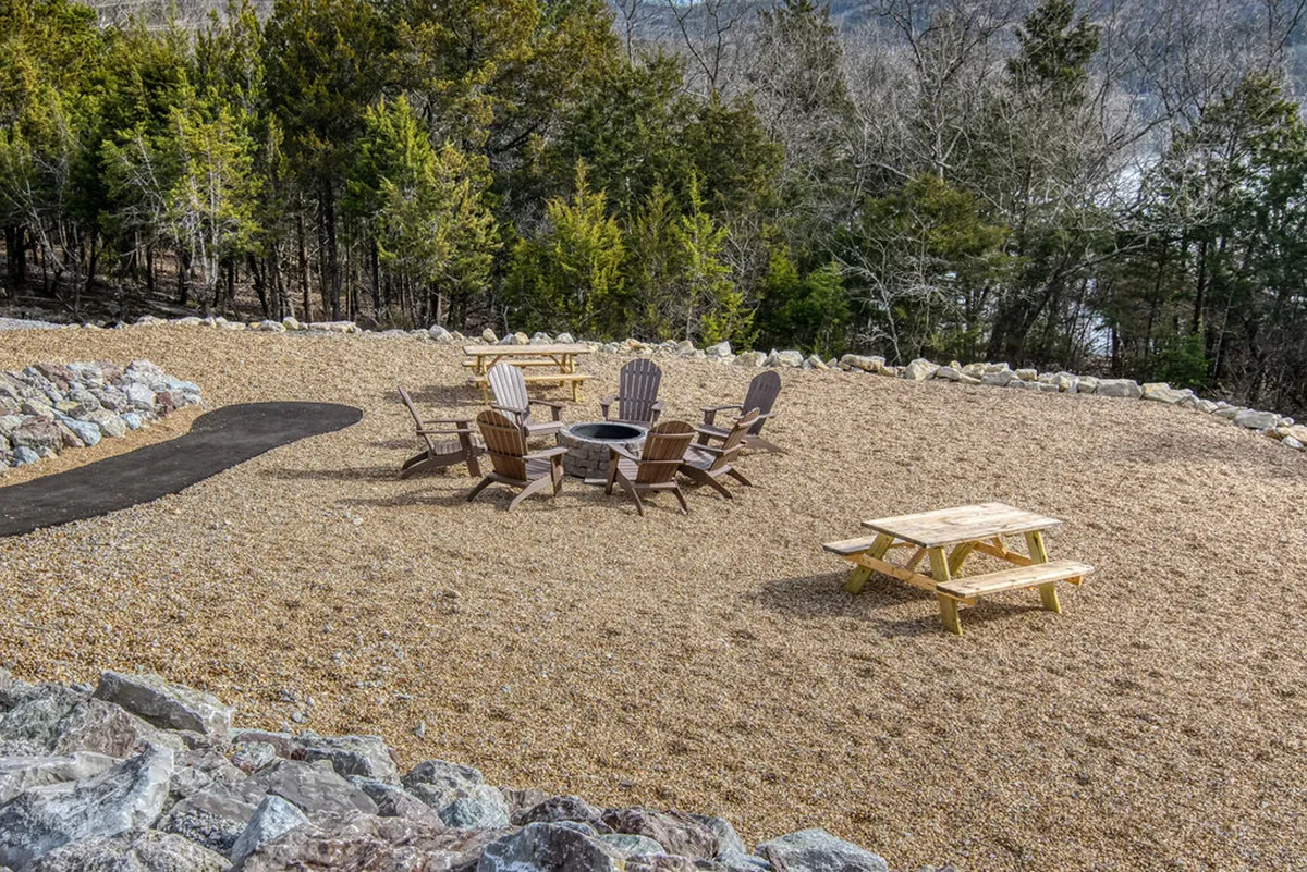 A relaxed evening scene around a communal fire pit, with adirondack chairs and a picnic table set beneath surrounding trees. The sun is setting, and guests are gathered around the fire sharing stories, marshmallows ready for toasting.