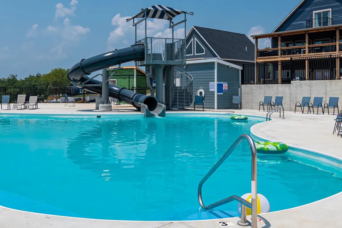 A lively outdoor pool scene with a water slide, green pool floats, and lounge chairs lining the pool deck. The area is bustling with activity as people enjoy swimming, sliding, sunbathing, and relaxing with friends under the warm sunset light.