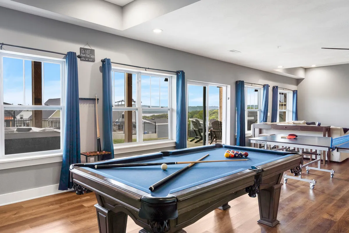 This image showcases a lively game room featuring both a pool table and a ping pong table set against large windows with an outdoor view. The space is designed for socializing, casual competition, and relaxation, with ample natural light creating a cheerful atmosphere.