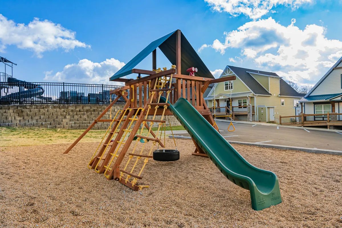 This exterior image centers on a modern playground structure with a slide and swing set, surrounded by a sand area. Children are actively playing, while families and parents gather nearby, enjoying outdoor social time.