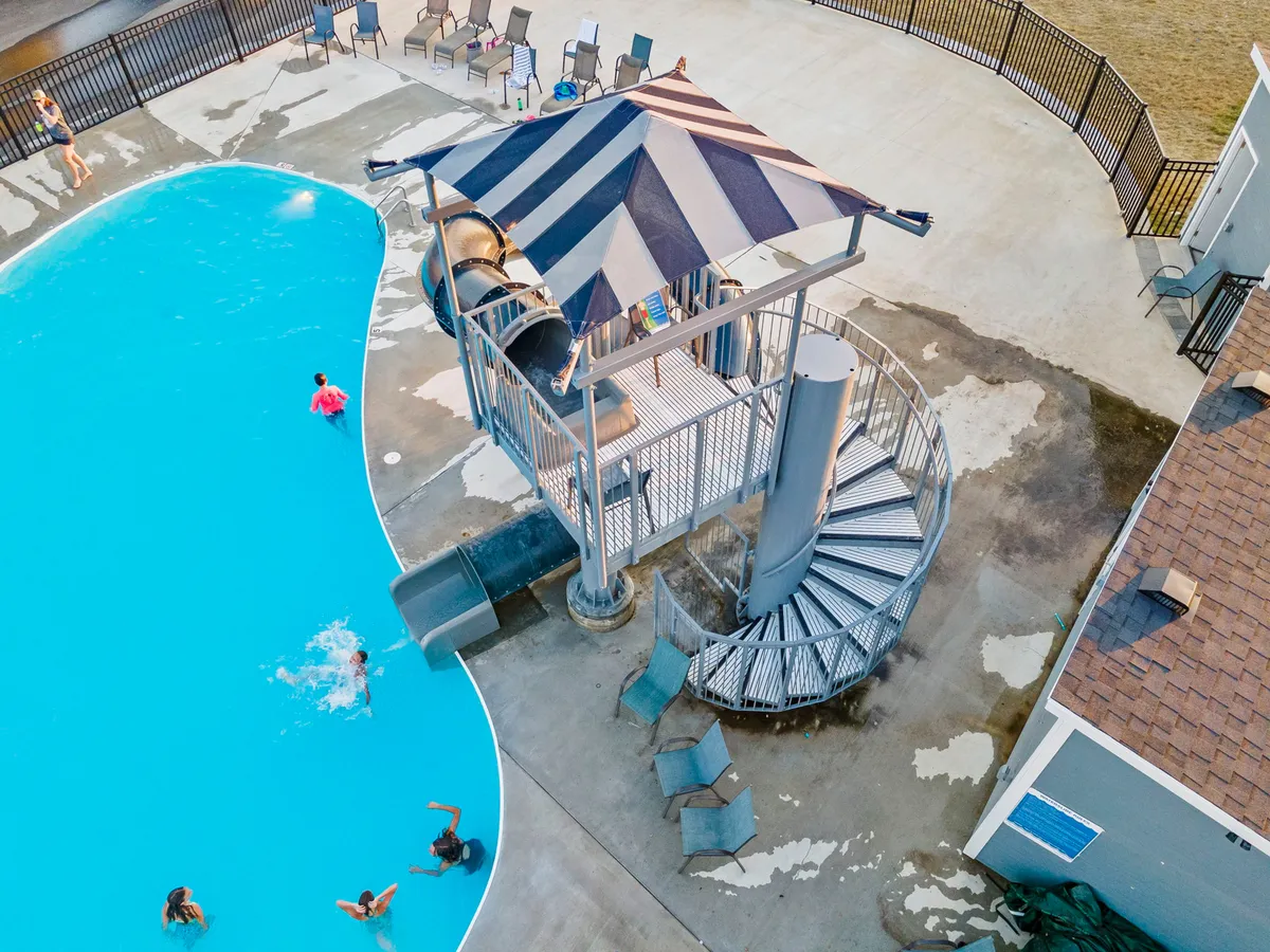 This lively exterior image showcases the outdoor swimming pool, complete with a vibrant blue water slide, loungers around the pool, and children engaging in splashy summer fun. Adults relax nearby, creating a dynamic atmosphere of multigenerational enjoyment.
